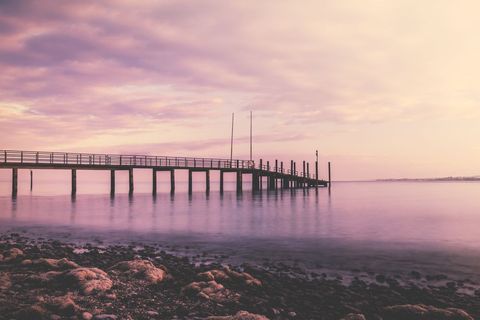 Pastel Sunset Pier Stretching Over Calm Sea Reflecting Pink Sky with Rocky Shore
