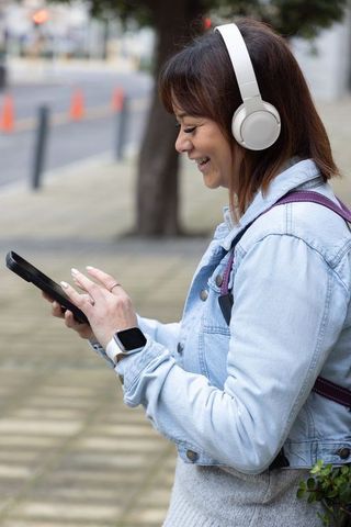 Asian woman smiling, tapping phone on city sidewalk wearing headphones and smartwatch