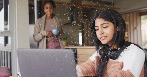 Teen girl studying on laptop with headphones while parent looks on in sunlit kitchen