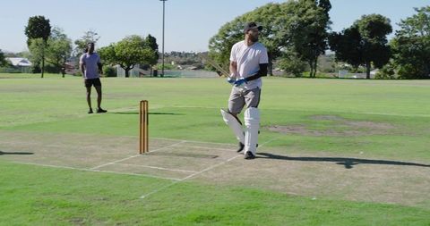 Cricketer batting at crease on sunny park pitch wearing pads gloves cap with wooden bat