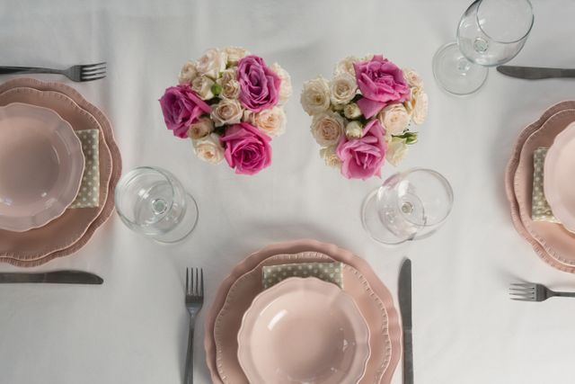 Overhead view of an elegant table setting featuring pink and white roses in floral arrangements. The table includes pink dinnerware, glassware, and cutlery, perfect for a special occasion or celebration. Ideal for use in event planning, wedding inspiration, dining decor, and romantic dinner themes.