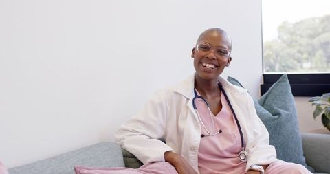Smiling female clinician wearing pink scrubs and white coat on sofa, welcoming portrait
