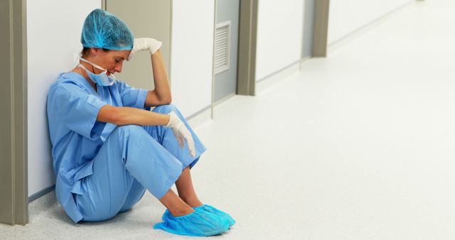 Nurse wearing scrubs and personal protective equipment, sitting on hospital floor in distress. Tired healthcare worker reflects high stress and demands of medical profession. Ideal for use in articles about medical professions, stress, healthcare challenges, emotional health in medical fields, and hospital settings.