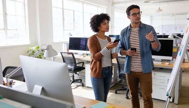 Two coworkers brainstorming at whiteboard in modern open-plan office holding smartphones