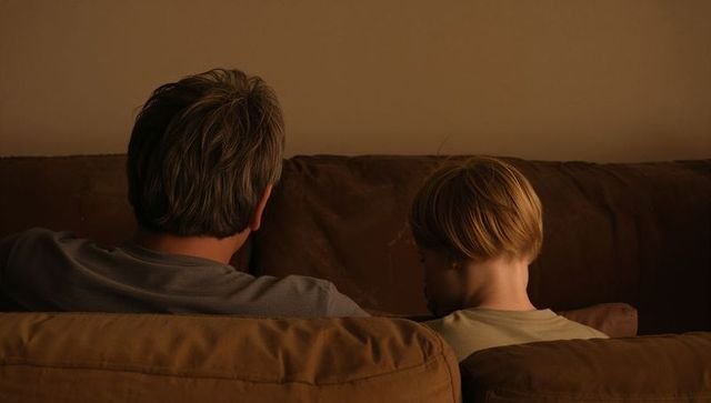 Adult and child sharing quiet evening on worn brown couch, warm low light, intimate moment