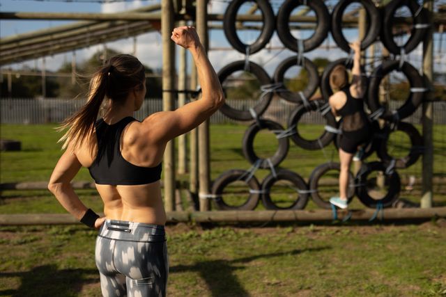Rear view of a Caucasian woman wearing sports clothes raising her fist and motivating another woman climbing over a wall of tyres on a climbing frame at an outdoor gym during a bootcamp training session