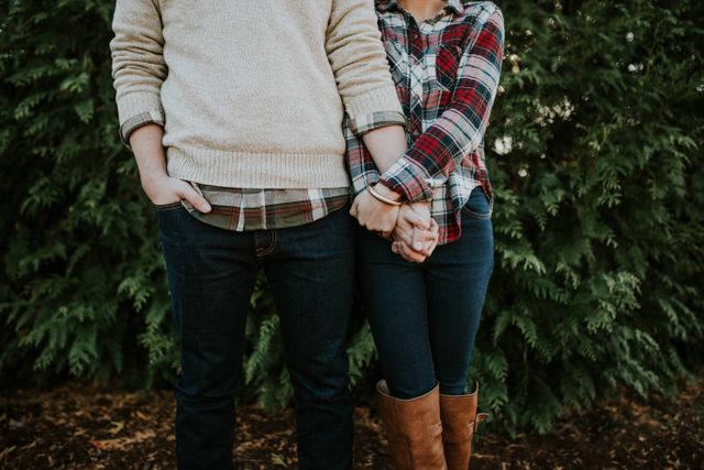 Couple holding hands while standing outdoors in casual fall outfits, featuring a plaid shirt and a sweater. The background highlights lush greenery, hinting at a nature or forest setting. Ideal for use in marketing materials related to relationships, love, fall fashion, outdoor activities, or lifestyle blogs.