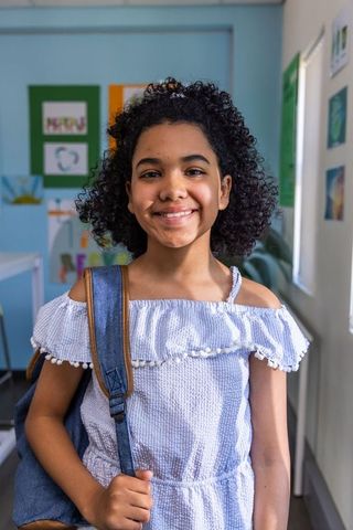 Smiling african american preteen wearing blue ruffle top holding denim backpack in classroom