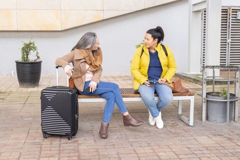 Mature women chatting on urban bench with luggage, casual travel conversation and friendship