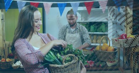 Smiling woman holding wicker basket picking fresh greens at market while vendor arranging produce