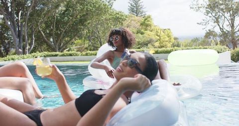 African american women relaxing on inflatable floats with lemon drinks in backyard pool