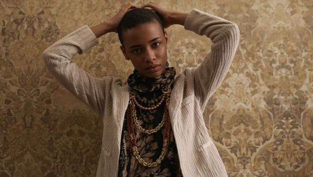 Edgy fashion portrait model with layered necklaces and beige blazer on ornate damask
