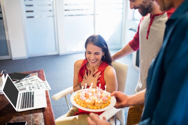 Colleagues are celebrating a birthday in the office. The scene includes a woman sitting in a chair, looking surprised and happy as a man presents a cake with candles. Another colleague stands nearby, adding to the cheerful atmosphere. Use this image for themes related to workplace culture, team bonding, and corporate events.
