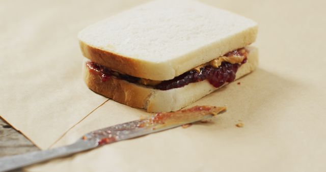 Homemade peanut butter and jelly sandwich resting on brown paper with knife featuring spread. Ideal for illustrating easy, classic snack options or lunch meals. Perfect for food blogs, recipe articles, children's lunch ideas, and culinary websites.