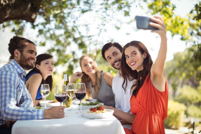Group of friends enjoying a meal at an outdoor restaurant, taking a selfie together. Perfect for use in advertisements for restaurants, social media campaigns, and lifestyle blogs. Highlights themes of friendship, leisure, and social gatherings.