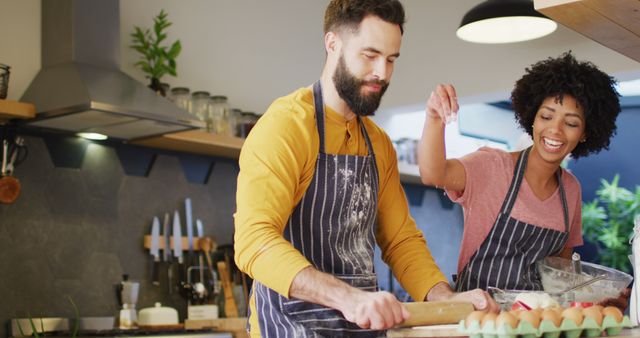 Couple dressed in aprons baking in a modern kitchen. The woman is smiling and adding ingredients while the man is rolling the dough, showcasing teamwork and enjoyment. Ideal for use in lifestyle, cooking blogs, home decor inspiration, culinary classes, relationship advice articles, or family-themed content.