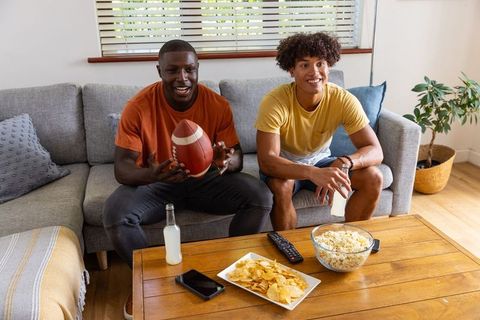 Black and mixed-race friends watching football at home holding ball and drinks