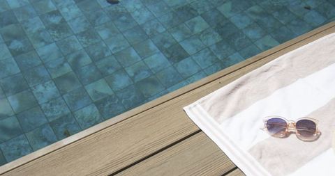 Sunlit wooden deck with striped towel and translucent sunglasses by blue tiled pool