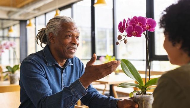 Smiling senior man offering potted magenta orchid in sunlit cafe