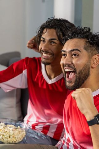 Friends cheering and fist-pumping in red and white jerseys while watching match on couch