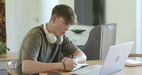 Teen studying at home with laptop and headphones writing notes at minimalist wooden table