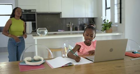 African american daughter doing homework with laptop while mother supervising in kitchen