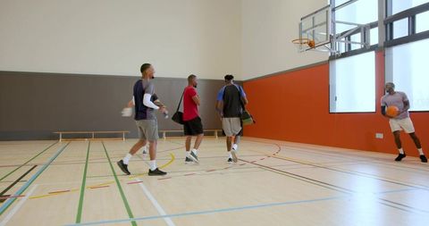 Diverse male basketball players walking into indoor court carrying balls and bags