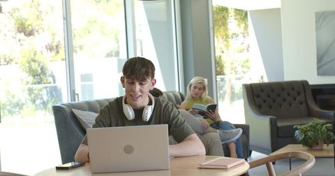 Teen male working on laptop with headphones in sunlit modern living room online learning