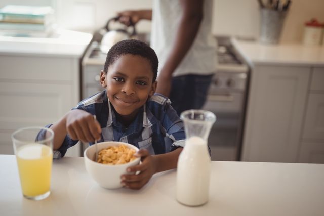 Young boy enjoying breakfast cereal at kitchen counter, smiling at camera. Glass of juice and milk jug on counter. Ideal for family lifestyle, morning routine, healthy eating, and domestic life themes.