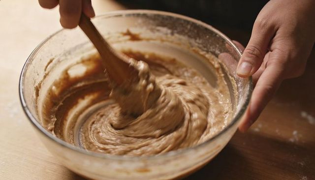 Hands stirring spiced brown batter in glass bowl using wooden spoon for baking