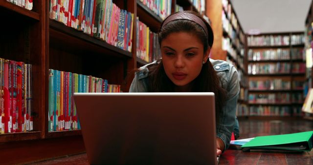 Young female student using laptop in library for studying and research. Suitable for use in content related to education, academic resources, technology in learning, and student life. Ideal for illustrating library environments, online learning, and dedication to studies.