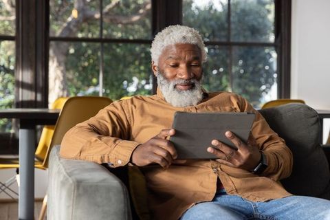 Senior man relaxing on couch using tablet and smartwatch beside sunlit window, cozy tech