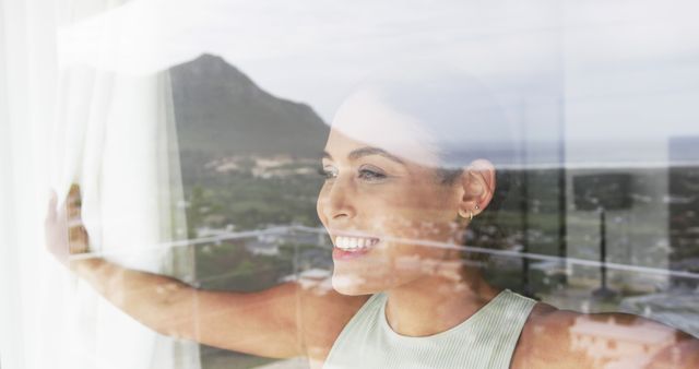 Young woman smiling while looking out of a window with a beautiful mountain view. Ideal for conveying themes of relaxation, peaceful retreat, connection with nature, and mental well-being. Suitable for blogs or articles on self-care, travel advertisements, wellness promotions, and lifestyle imagery.