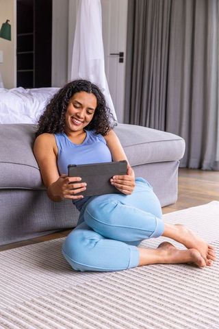 Smiling woman relaxing on bedroom floor using tablet by gray sofa