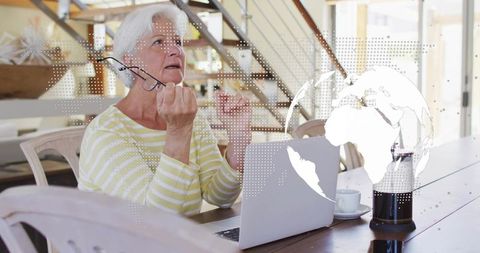 Senior woman talking on video call holding glasses at laptop with coffee and globe overlay