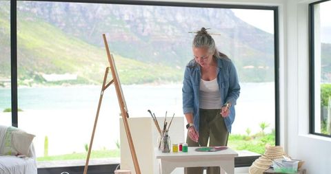 Mature woman painting in bright coastal studio with easel and waterfront view