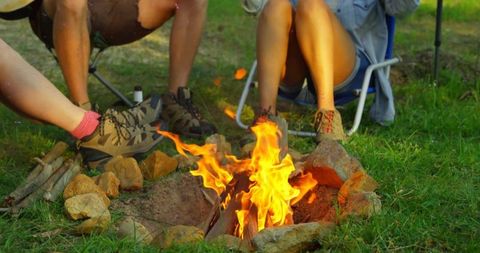 Group warming feet by campfire in meadow relaxing on folding chairs during sunny camping