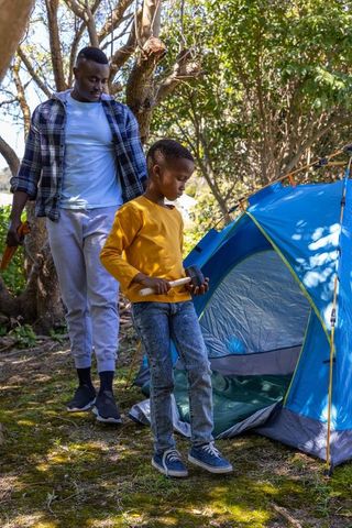 African american father and son pitching blue tent in forest during daytime camping