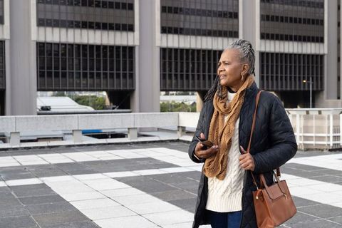 Senior african american woman standing on urban terrace holding phone, carrying leather bag