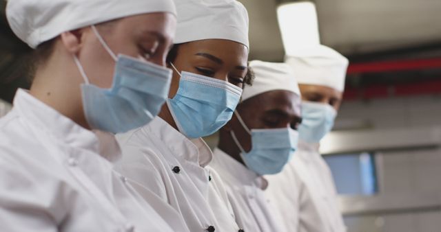 Chefs standing in a professional kitchen, wearing white uniforms and face masks, concentrating on their tasks. Ideal for use in articles, blogs, and marketing materials related to the culinary industry, hygiene standards in restaurants, or teamwork in professional kitchens.