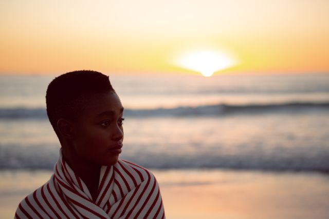 Woman standing on beach wrapped in blanket during sunset, looking thoughtful. Ideal for themes of contemplation, solitude, and peaceful moments. Suitable for use in articles, blogs, and advertisements focusing on relaxation, nature, and mental well-being.
