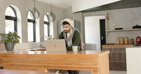 Indian man working remotely at modern wooden kitchen island with laptop and coffee mug