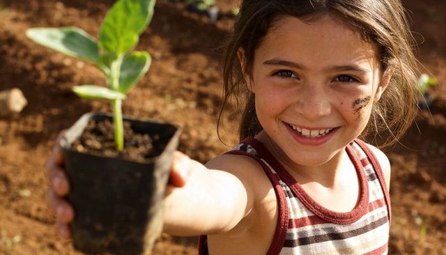 Smiling girl offering seedling in pot, learning gardening and sustainability outdoors