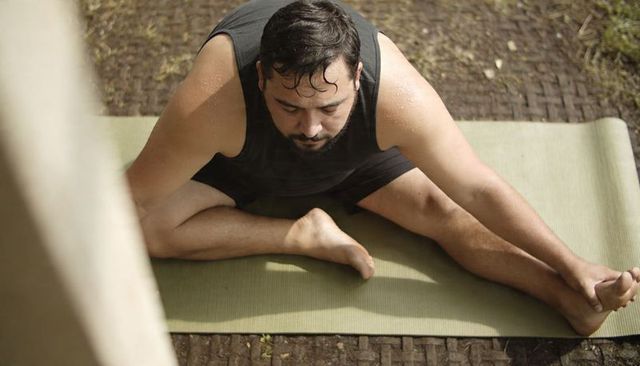 Adult man stretching on green yoga mat outdoors on brick patio, wearing black shorts