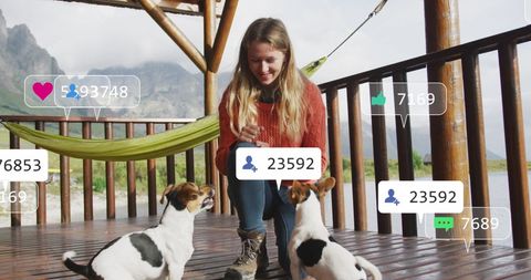 Young woman interacting with two dogs on a wooden terrace overlooking a scenic lake. Social media engagement emoticons and counters appear above, indicating online interactions. Ideal for illustrating themes around outdoor communication, digital connectivity, relaxed leisure time, and online viral content.