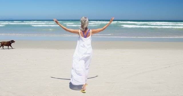 An elderly woman in a white dress is stretching her arms wide, embracing a peaceful moment on a sandy beach. Ocean waves roll gently towards the shore, creating a tranquil backdrop. The woman and her environment evoke a feeling of relaxation and carefree leisure. A dog is strolling in the background, adding a touch of life and companionship. This image can be used for themes including retirement, peace, self-care, summer vacations, beach activities, and personal wellbeing.