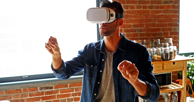 Man wearing a VR headset and denim shirt, standing in front of brick wall, engaging with a virtual environment in a modern home. Ideal for advertisements related to virtual reality technology, innovative home entertainment solutions, tech gadgets, and modern lifestyle visuals.