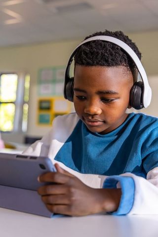 African american schoolboy concentrating on tablet wearing headphones in sunny classroom