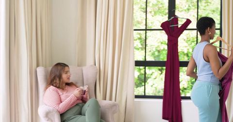 African american friends choosing burgundy dress by window while drinking coffee at home