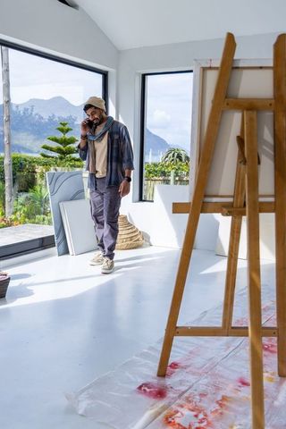 Artist talking on phone walking by sunny studio windows with easel and mountain view
